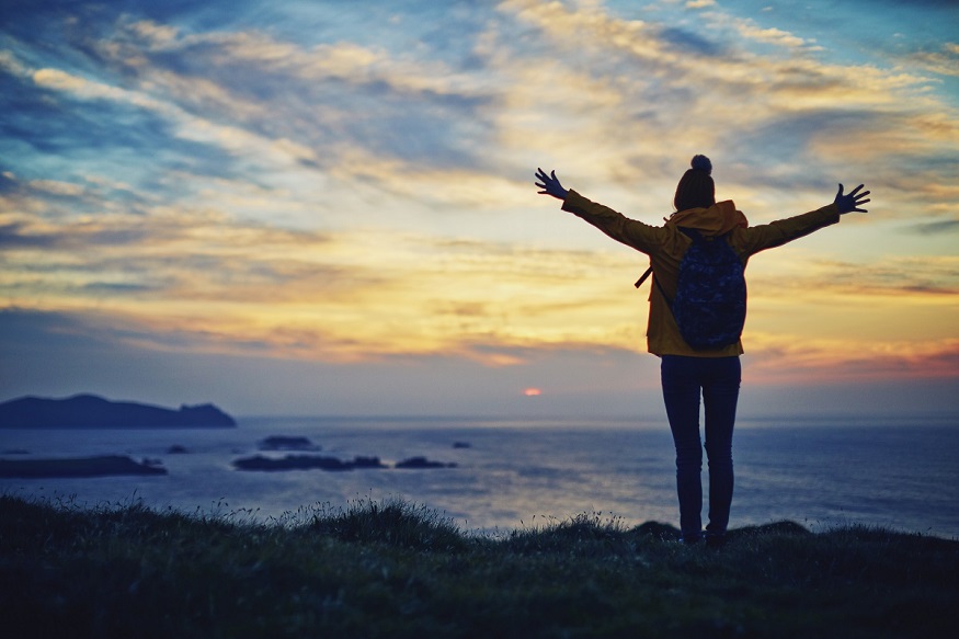 Carefree hiker standing with arms outstretched by sea during sunset